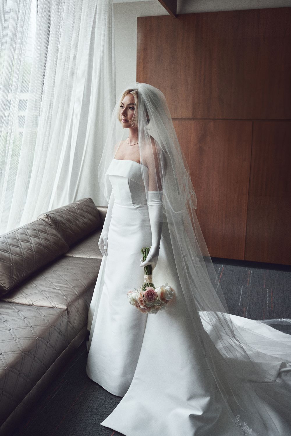 A bride in an elegant white wedding gown and veil poses gracefully by a window with sheer curtains and modern decor.