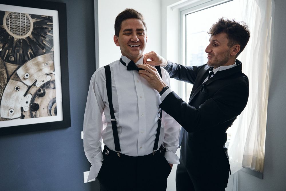 Two people in formal attire laugh and interact playfully in a bright room with white walls and windows.