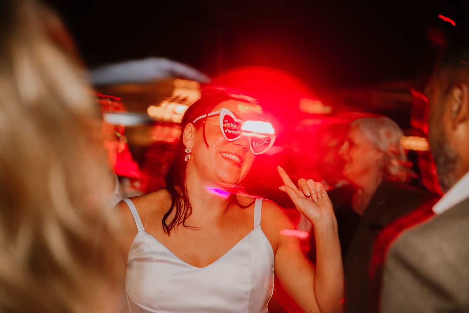 Bride dancing wearing heart-shaped glasses with red light streaming across her face, capturing a fun and artistic wedding reception moment.