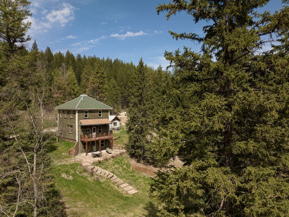 A rustic mountain cabin nestled among evergreen trees on a grassy hillside with blue skies overhead.