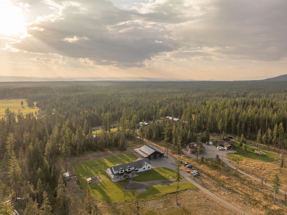 Aerial view of a rural property with buildings surrounded by vast evergreen forest and dramatic cloudy sunset sky.