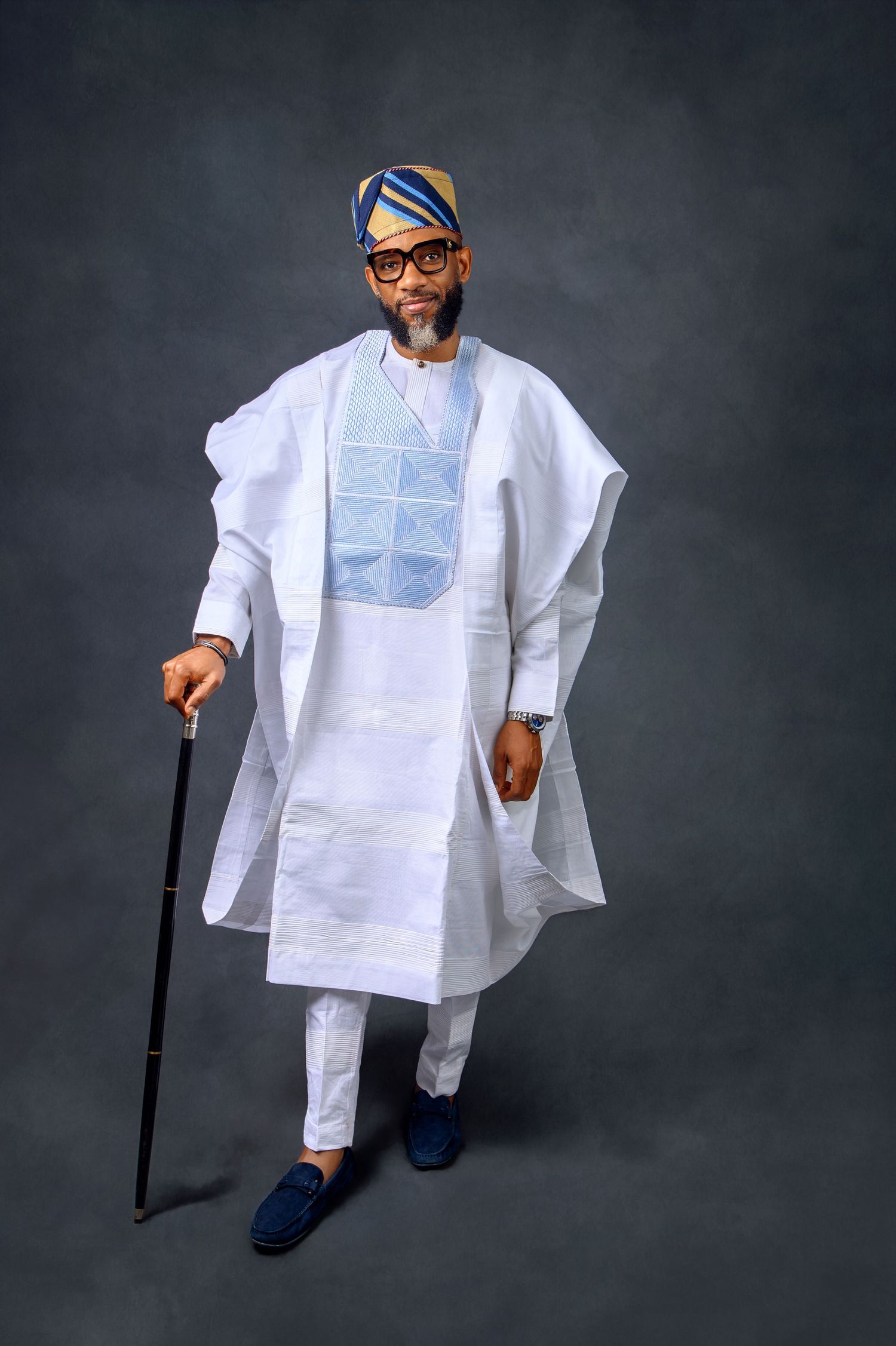 Traditional African agbada outfit in white with blue embroidery paired with blue suede shoes and walking stick.