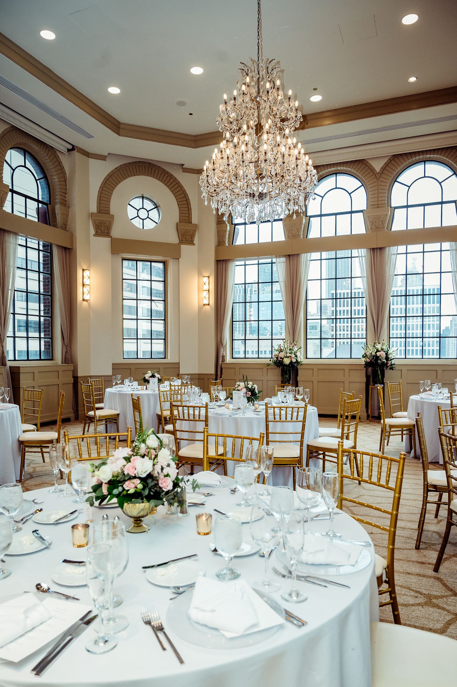 Elegant wedding reception venue with crystal chandelier, arched windows, and round tables set with white linens and gold chairs.