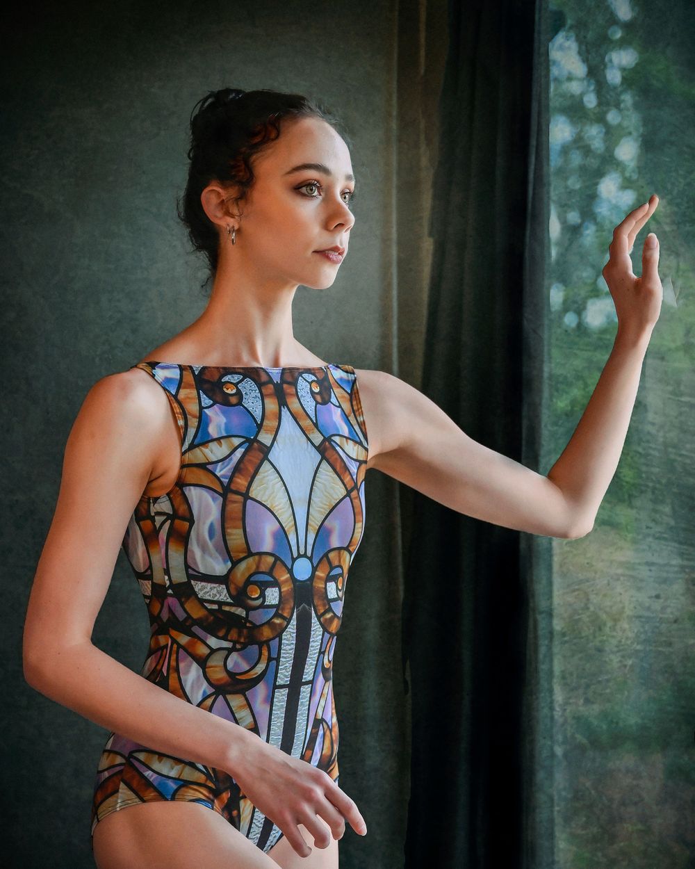 Dancer in stained glass pattern leotard poses gracefully by window with artistic lighting.