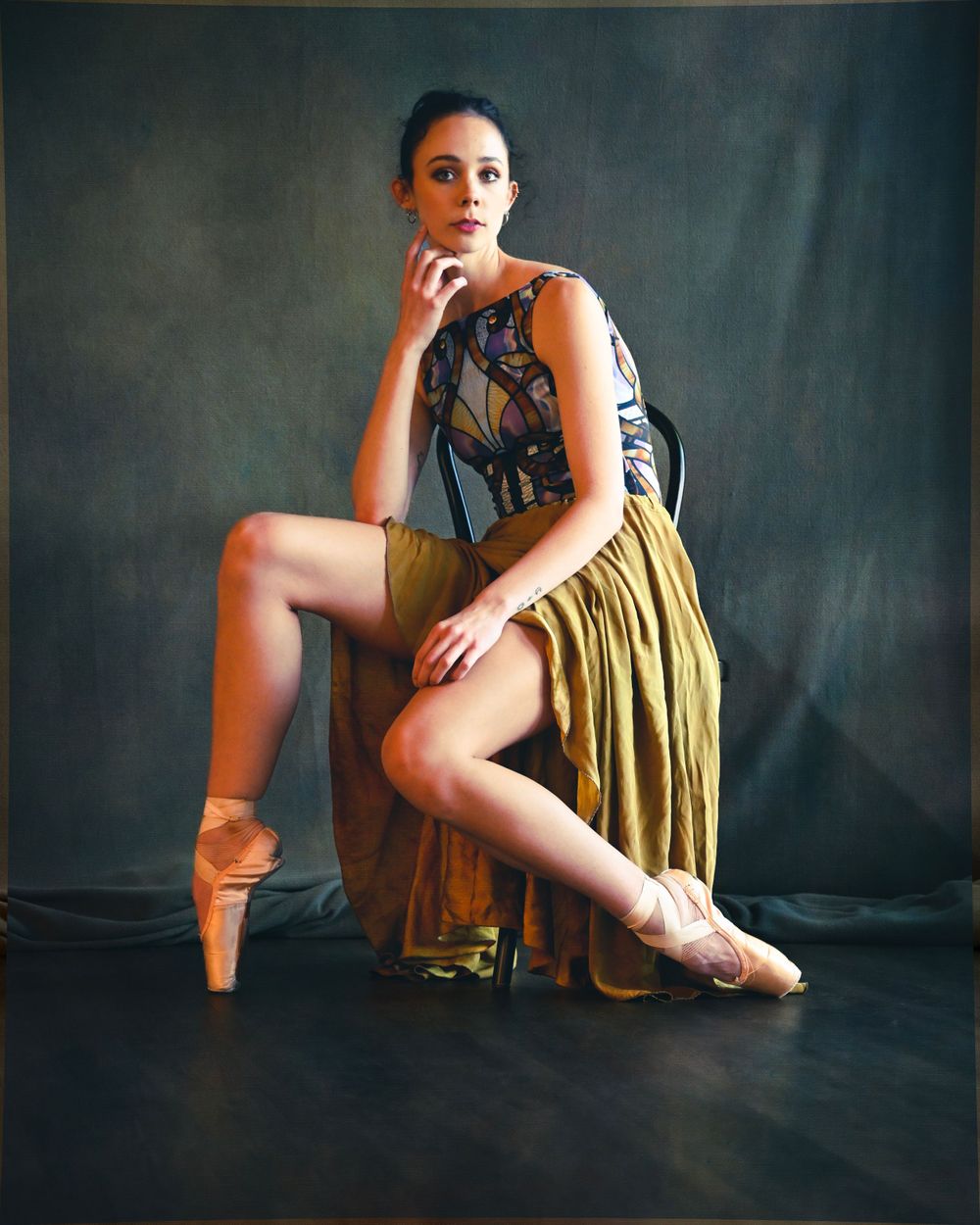 Ballerina poses in yellow skirt and patterned leotard against dark studio backdrop.