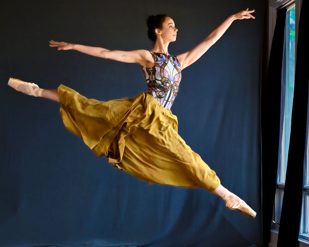 Dancer in yellow skirt and patterned top performs dramatic leap with arms outstretched.