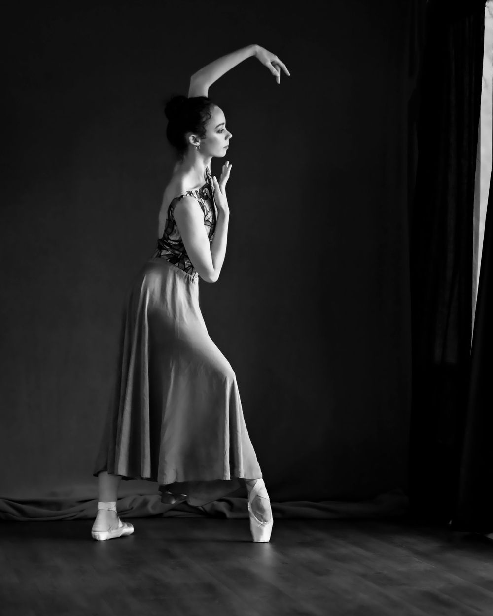 Ballet dancer in flowing dress strikes graceful pose in black and white studio photo.