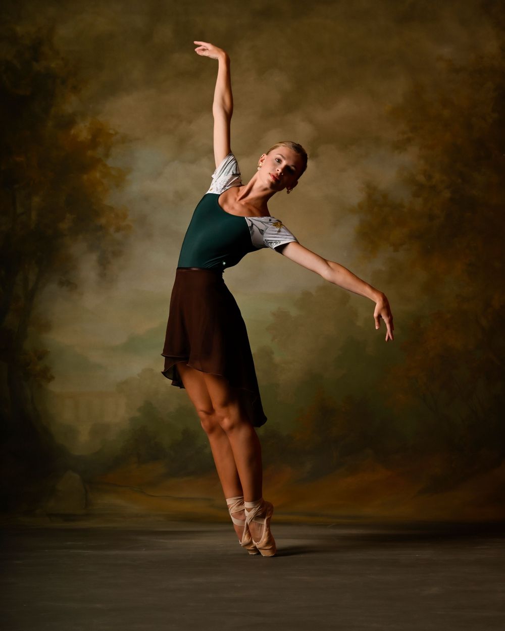 Ballet dancer in green and brown dress performs an elegant pose on point with dramatic lighting.