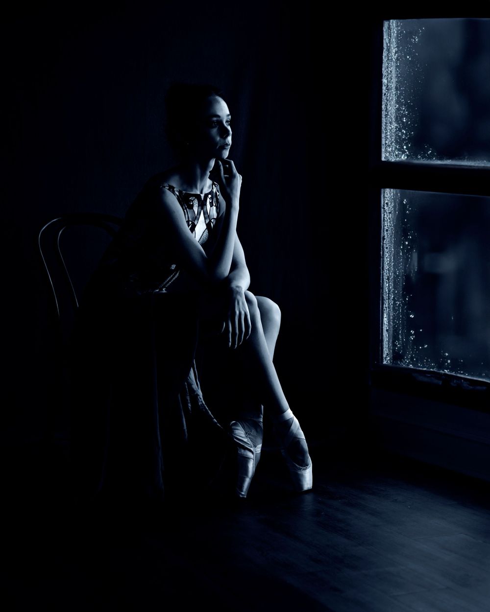 Dramatic low-key portrait of dancer by window in dark room with moody lighting.