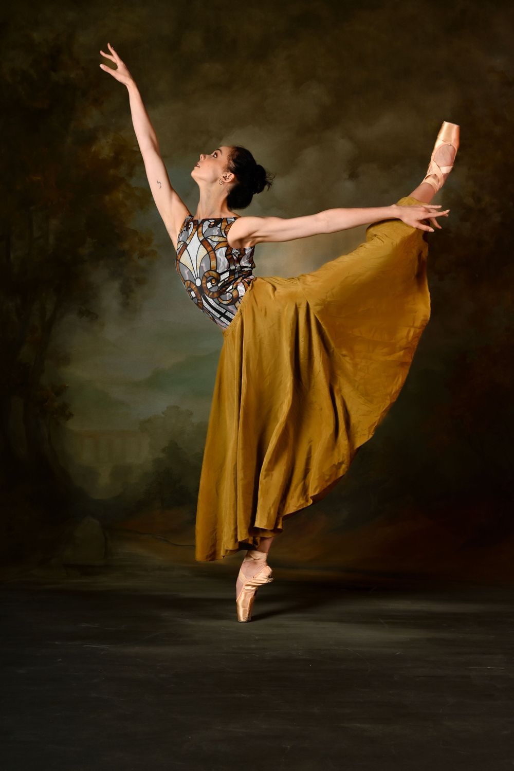 Dancer in gold skirt and patterned top strikes graceful arabesque pose against dark backdrop.
