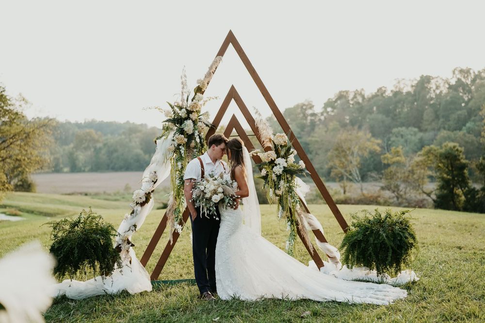 Newlyweds stand under triangular wooden arch decorated with greenery in scenic field.