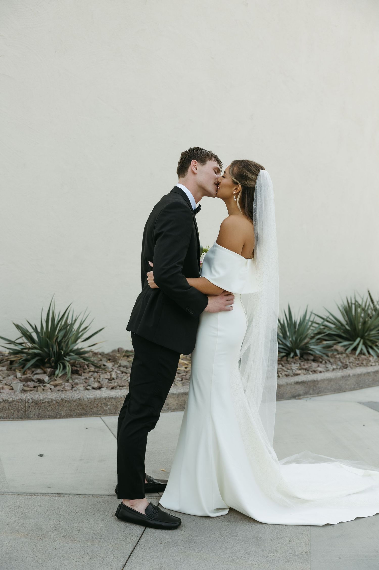 Romantic wedding kiss against minimalist white wall with desert landscaping.
