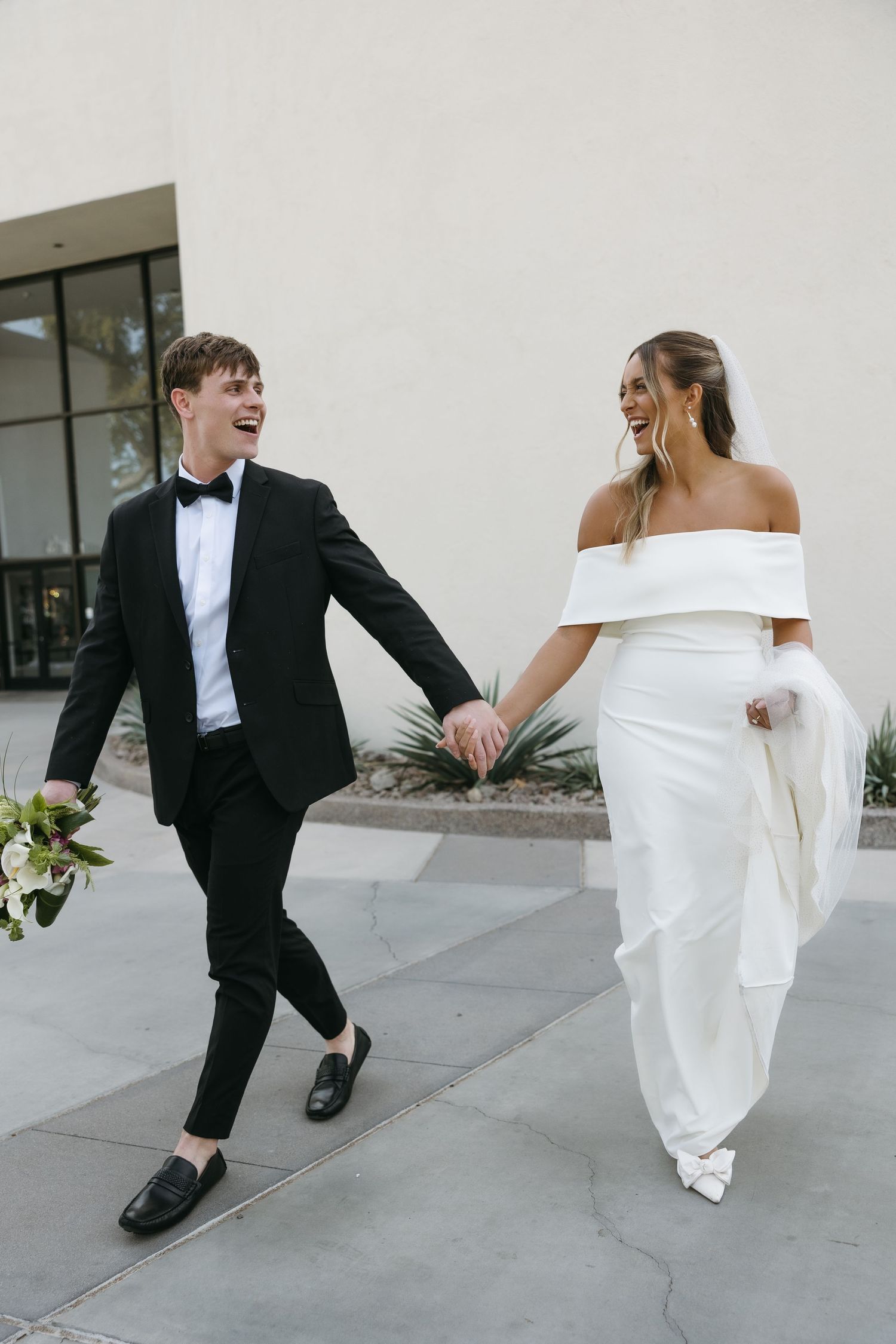 Joyful candid moment of newlyweds laughing and holding hands while walking outside.