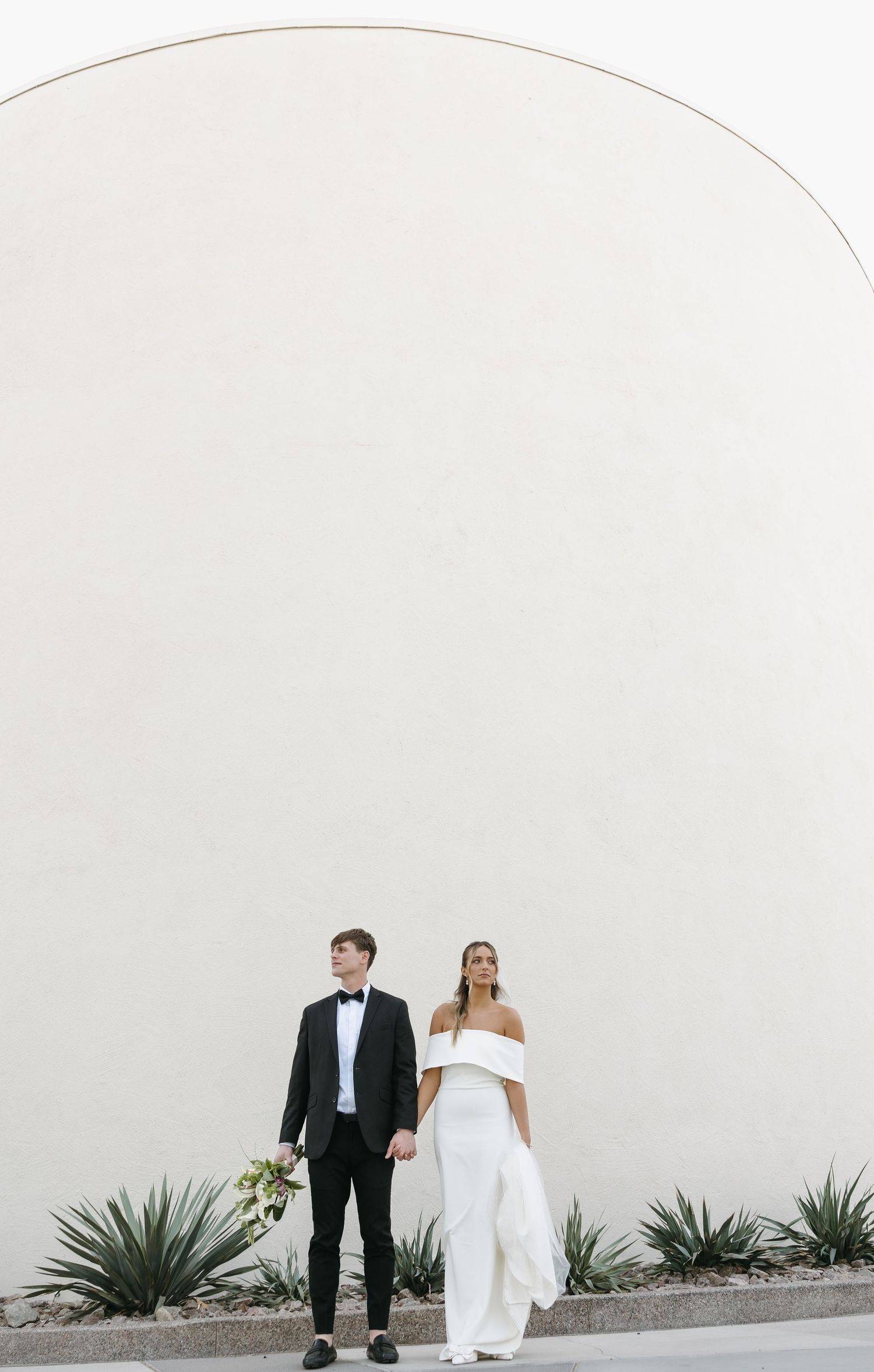 Minimalist wedding portrait of couple standing against white curved wall with desert plants.