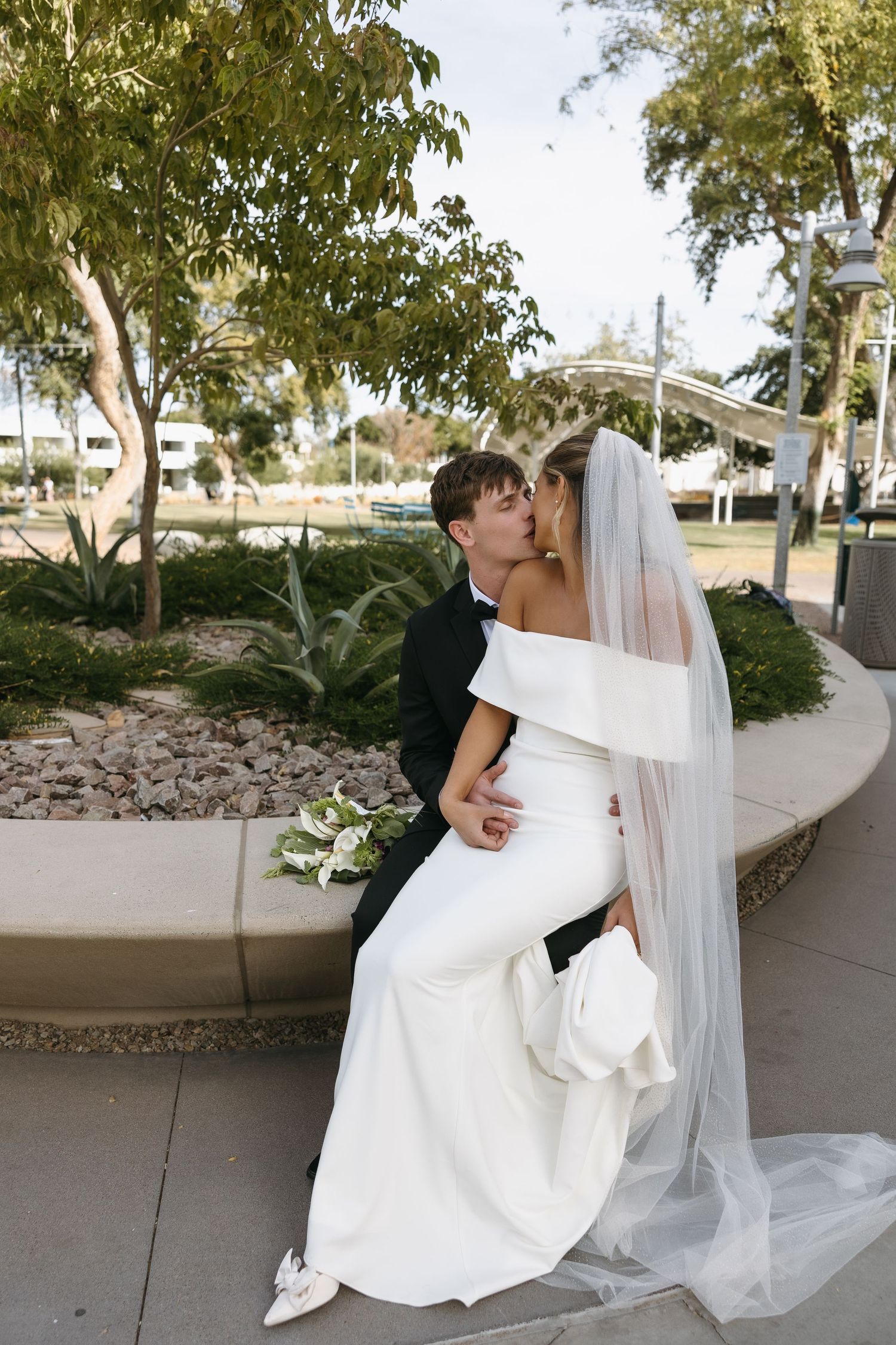 Romantic outdoor wedding photo of couple embracing on garden path with trees in background.