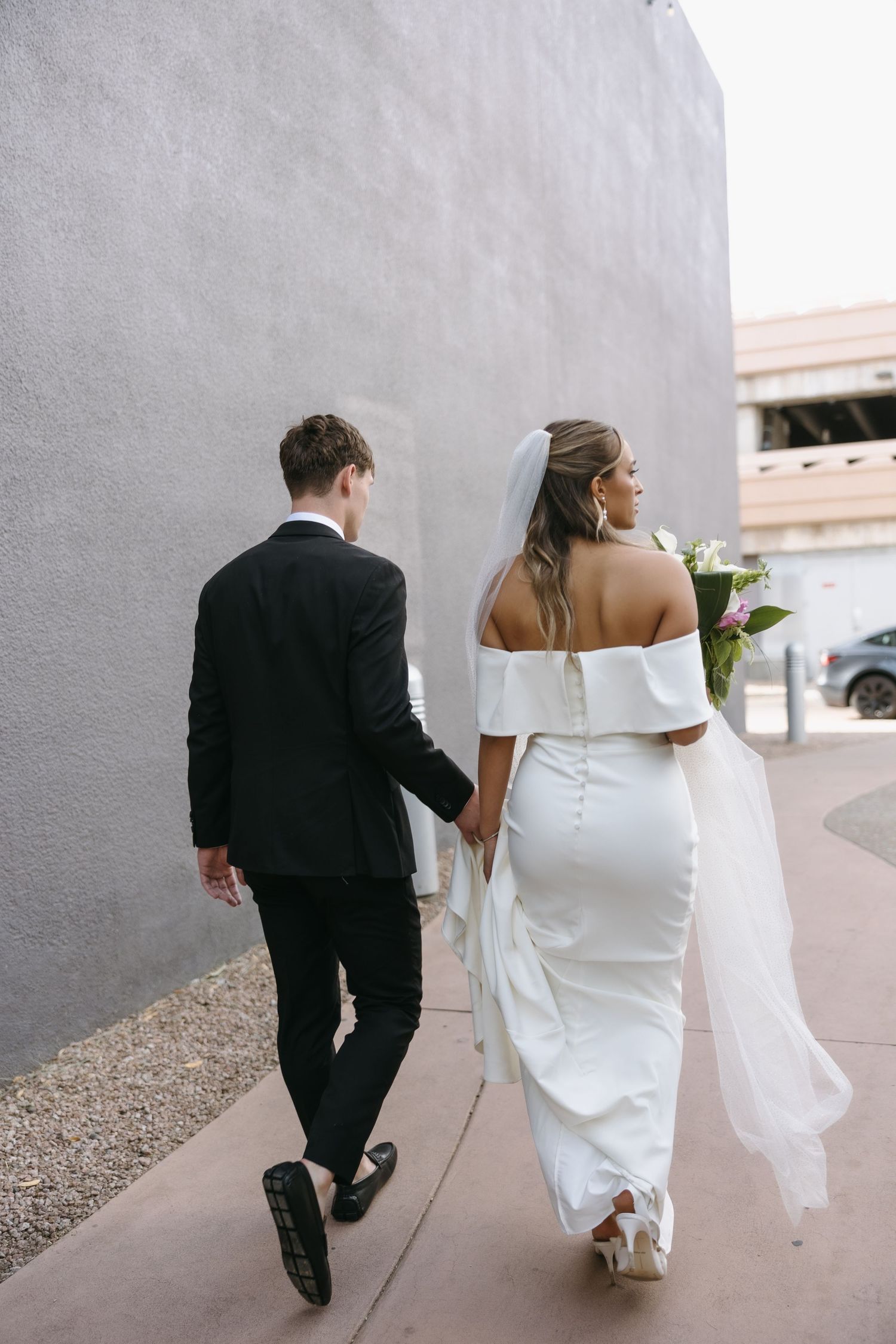 A bride in a white off-shoulder gown and groom in black suit walking together along a path.