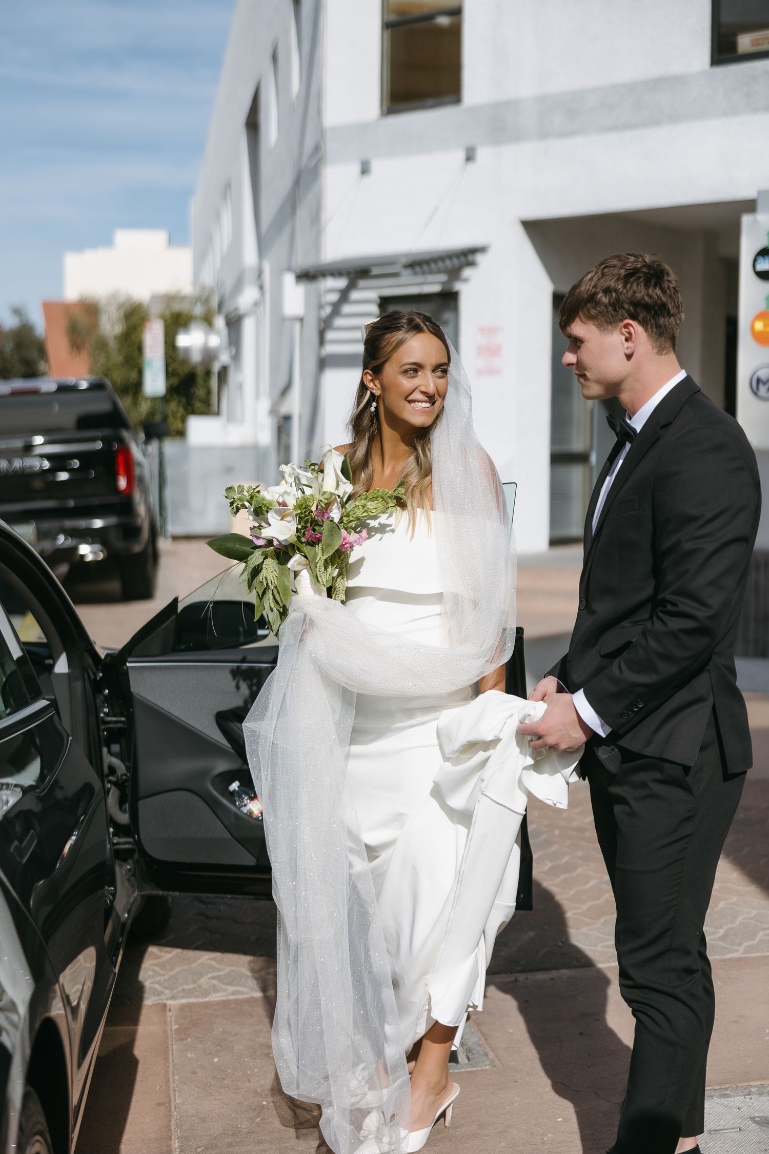 A couple walking along a white building on their wedding day with a floral bouquet.