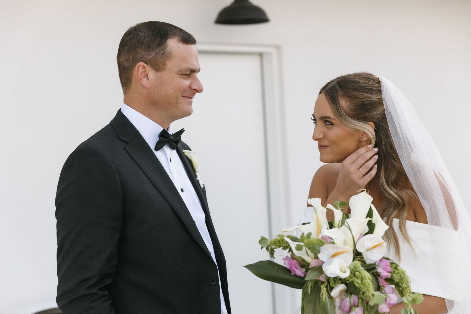 Tender wedding moment captured against white wall with elegant floral bouquet.