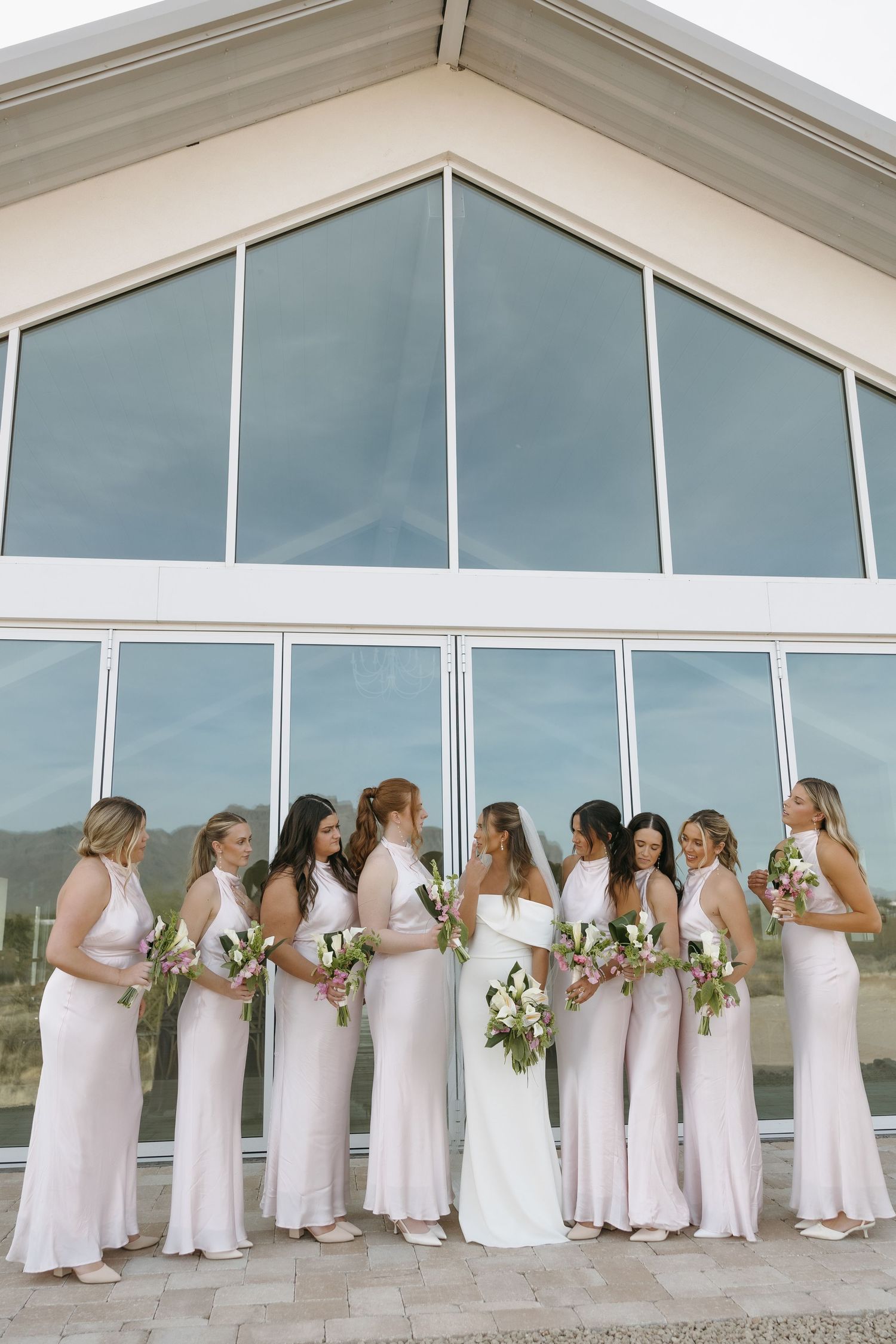 Wedding party in pink bridesmaid dresses poses in front of modern glass window exterior.