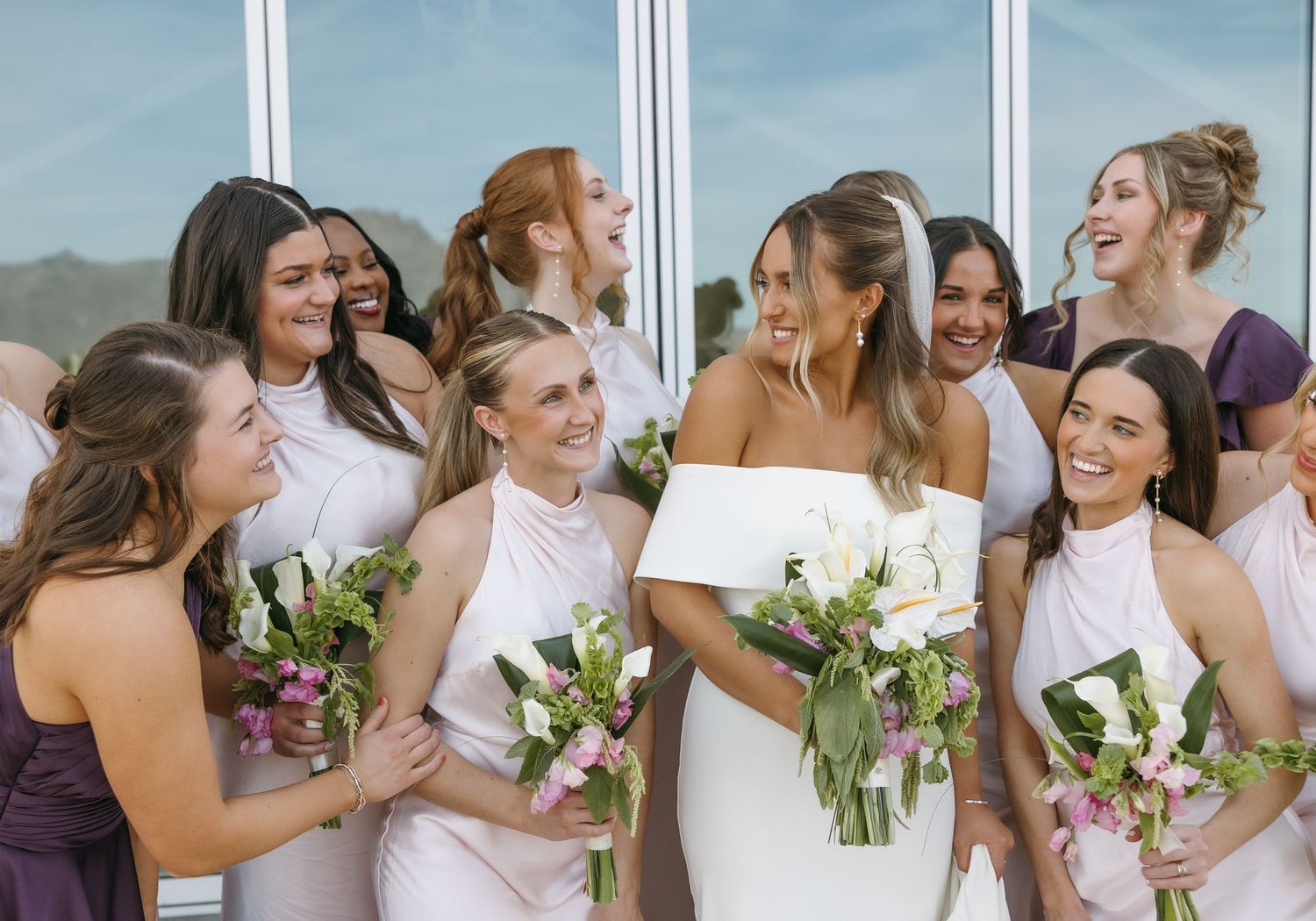 Bridal party in pink dresses sharing joyful moment with bouquets against glass backdrop.