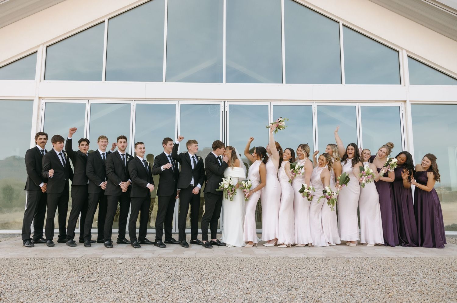 Large wedding party in formal attire lined up against modern glass building exterior.