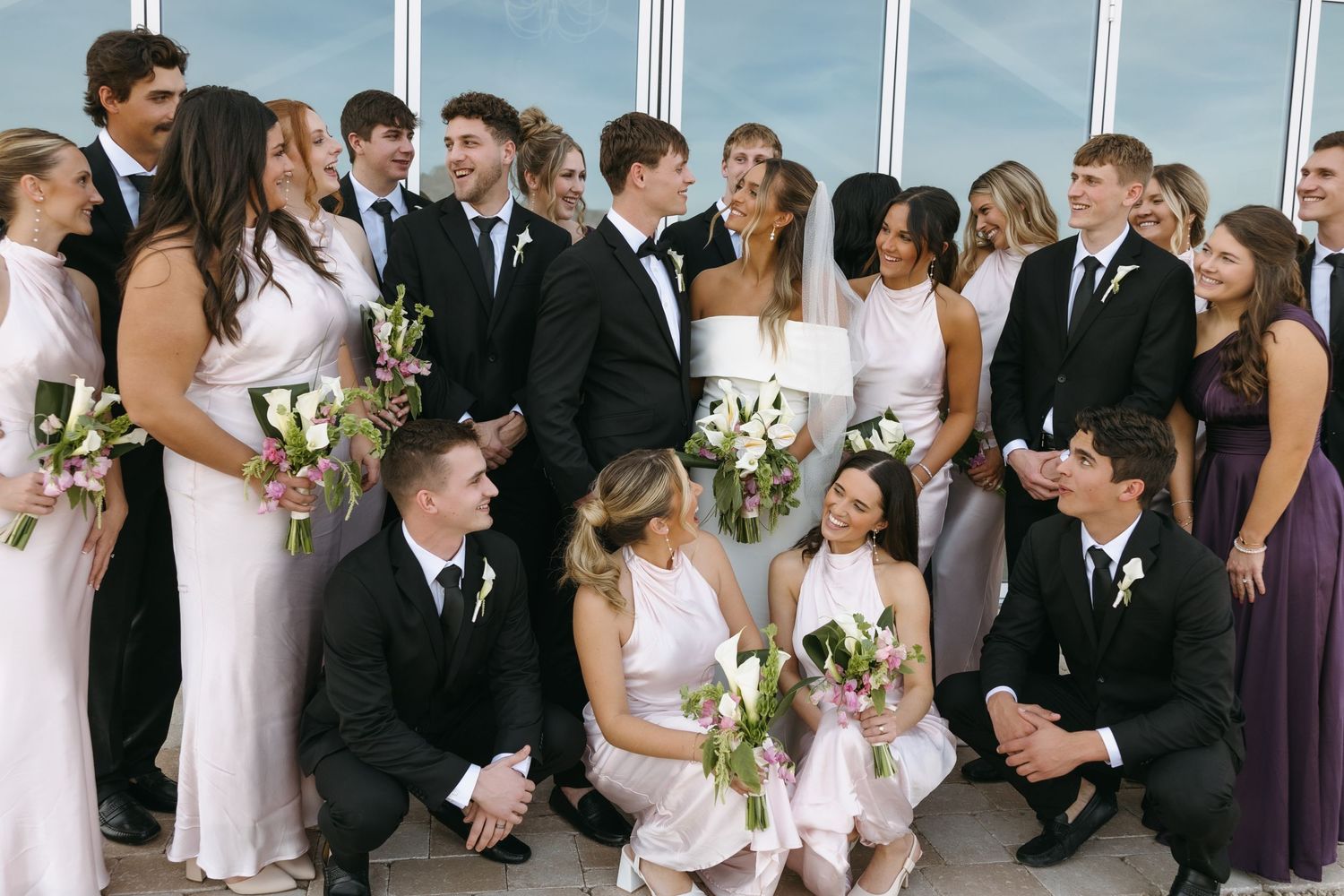 Wedding party gathered for casual group photo with flower girls in front of glass building.