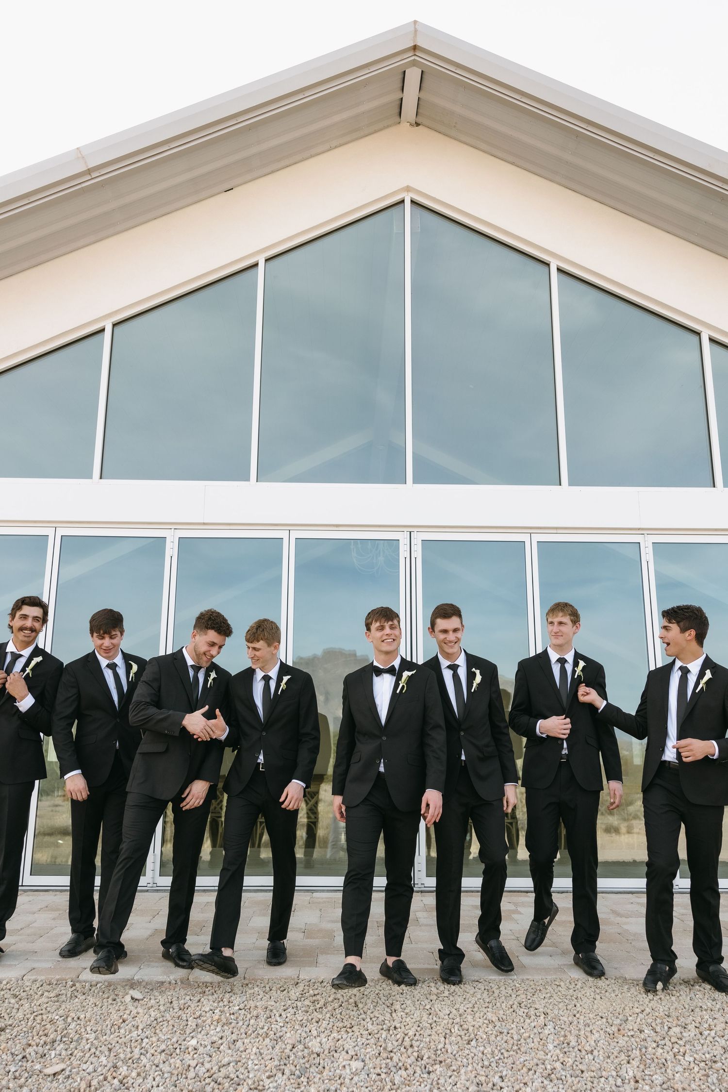 A group of groomsmen in black suits and ties pose together in front of a modern glass building.