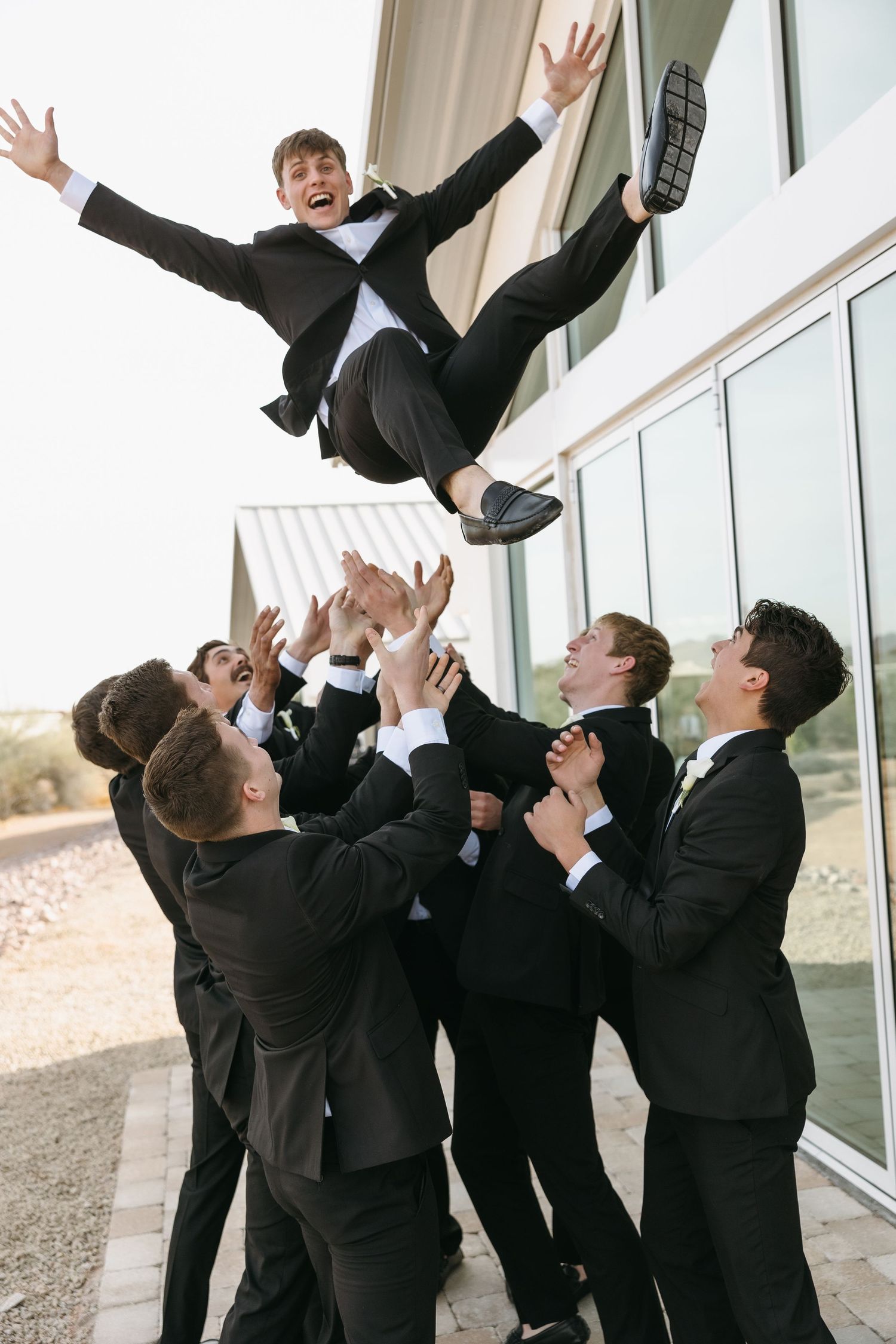 A fun wedding party photo of groomsmen tossing someone in the air outside a venue.