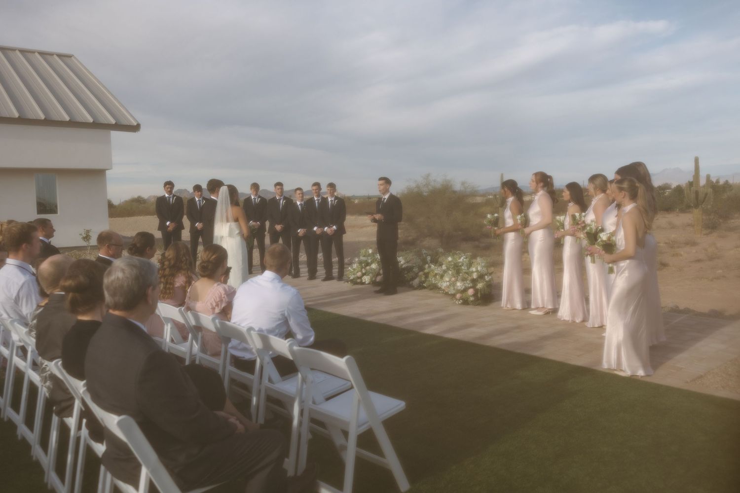 Wedding ceremony setup with white chairs on lawn and bridal party in elegant attire.