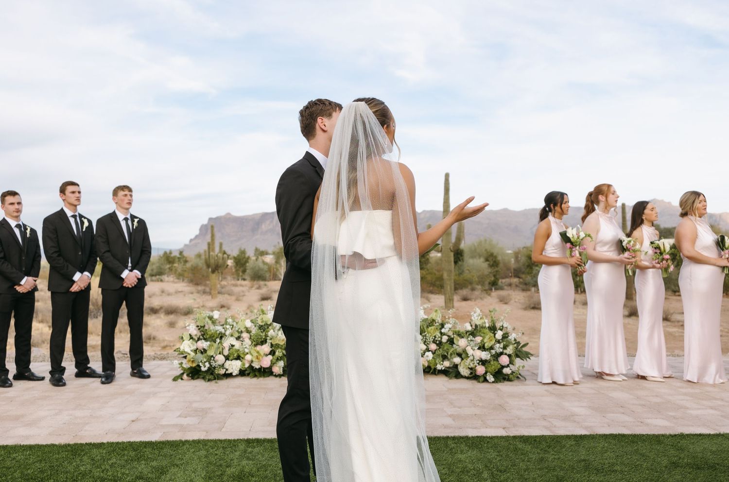 Wedding ceremony in desert setting with bridal party and floral arrangements.