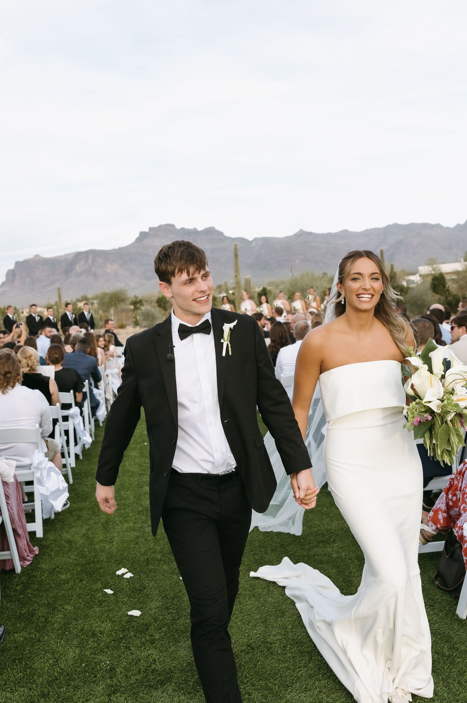 Newlyweds walk down aisle with desert mountains in background as guests look on.