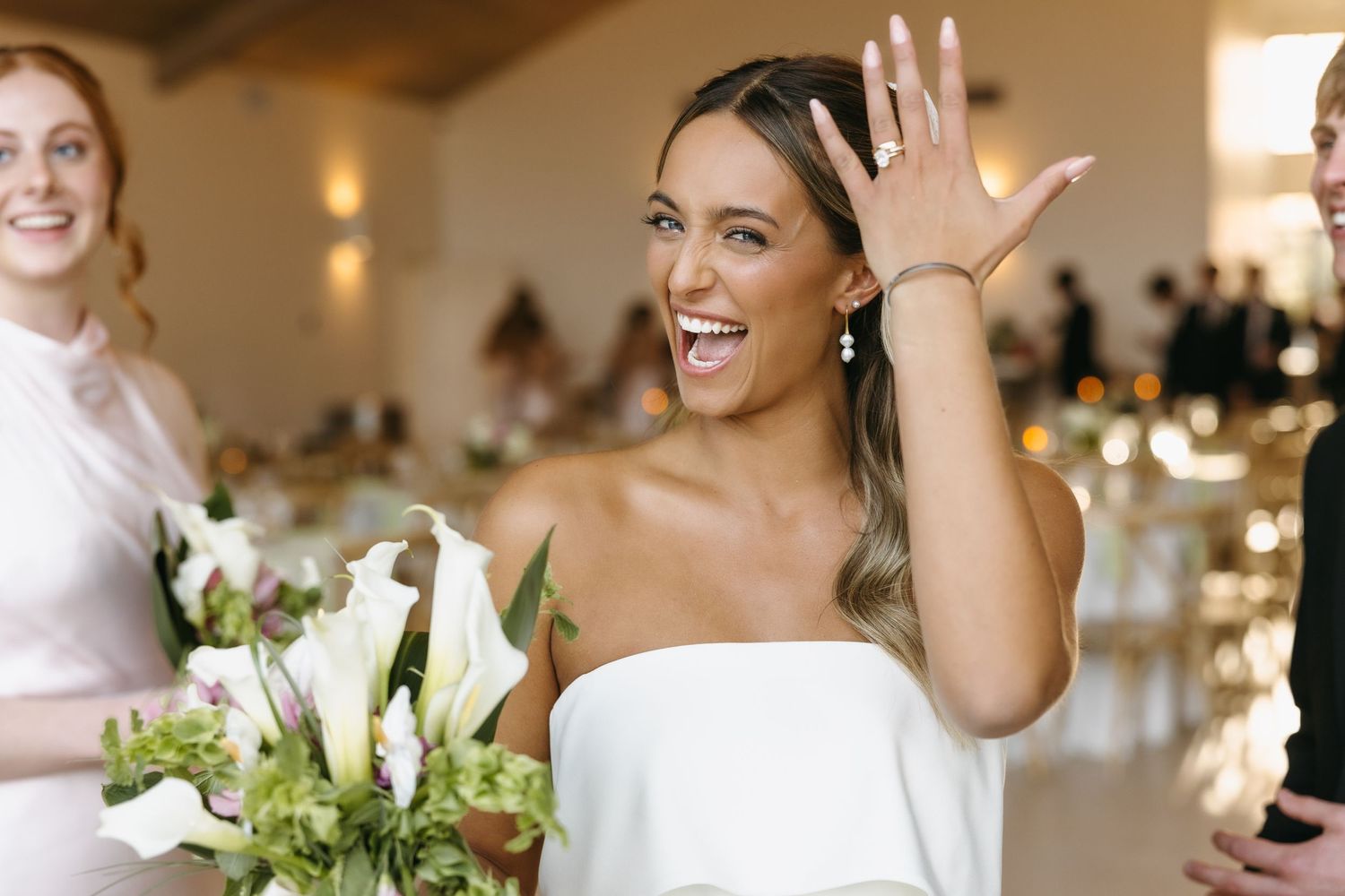 A joyful bride in a strapless white dress holds a bouquet of white flowers while waving at her wedding reception.
