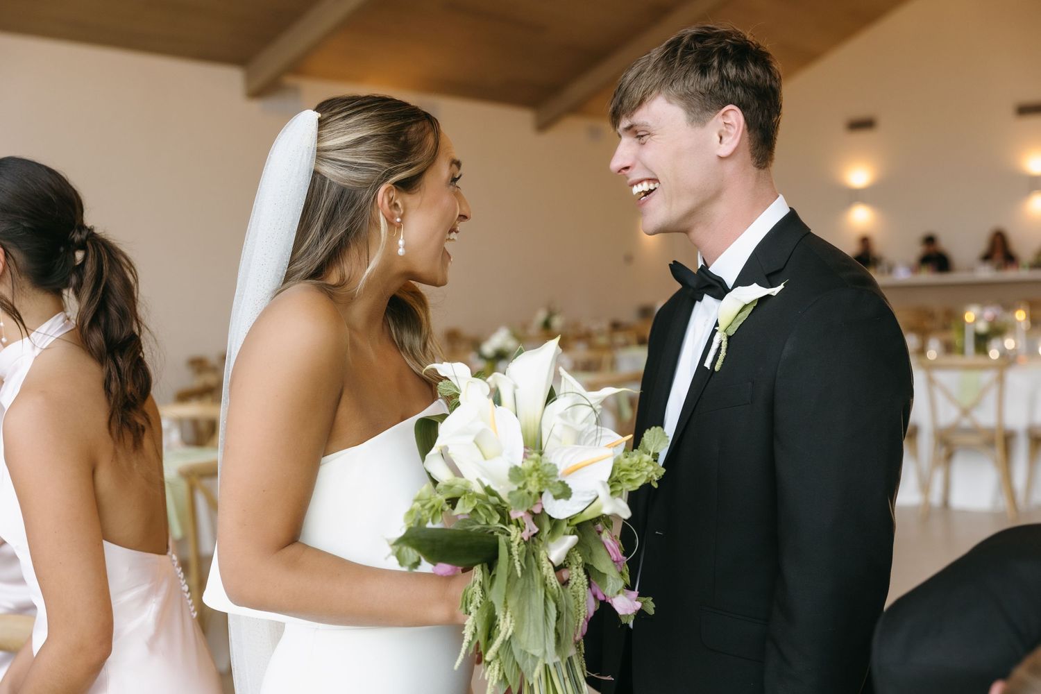A candid moment between newlyweds sharing laughter during their wedding ceremony with white calla lily bouquet.
