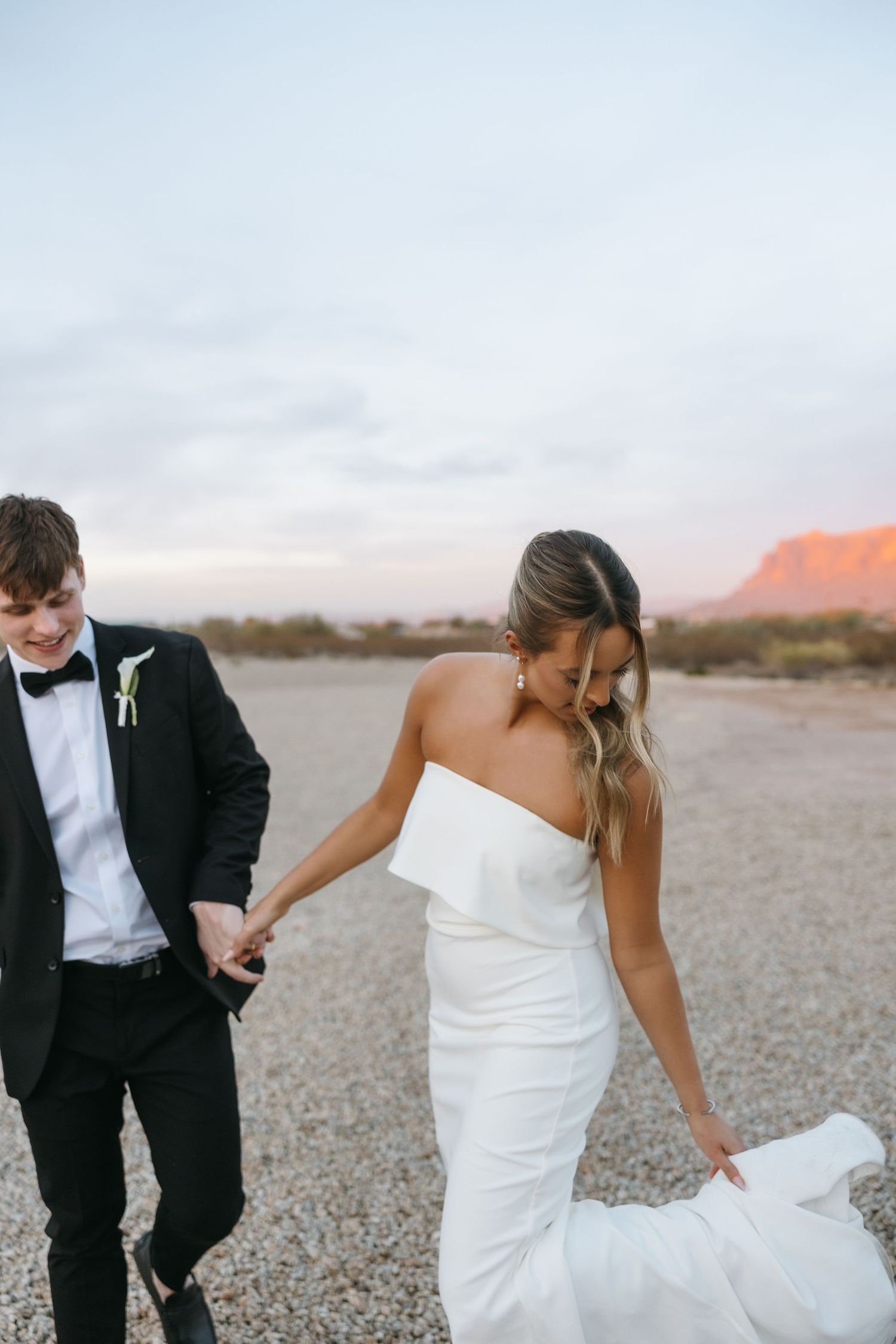 Couple in wedding attire walking through a desert landscape with mountains in the background.