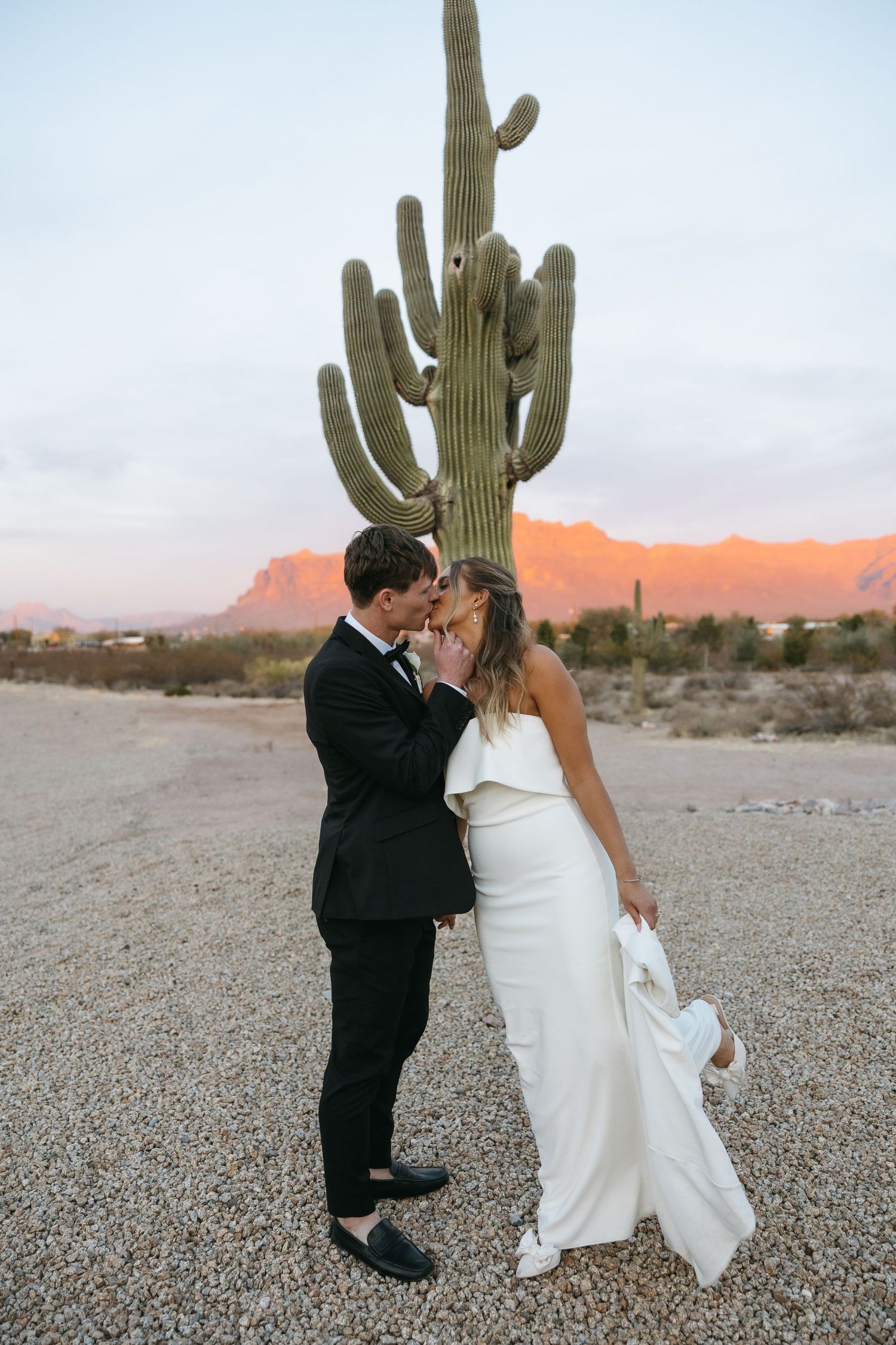 Romantic wedding portrait of couple embracing near a tall saguaro cactus in the desert at sunset.