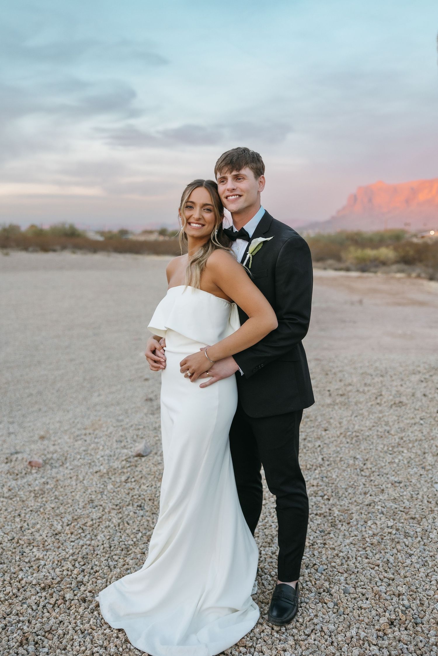 Desert wedding portrait with couple in formal attire against mountain backdrop at sunset.