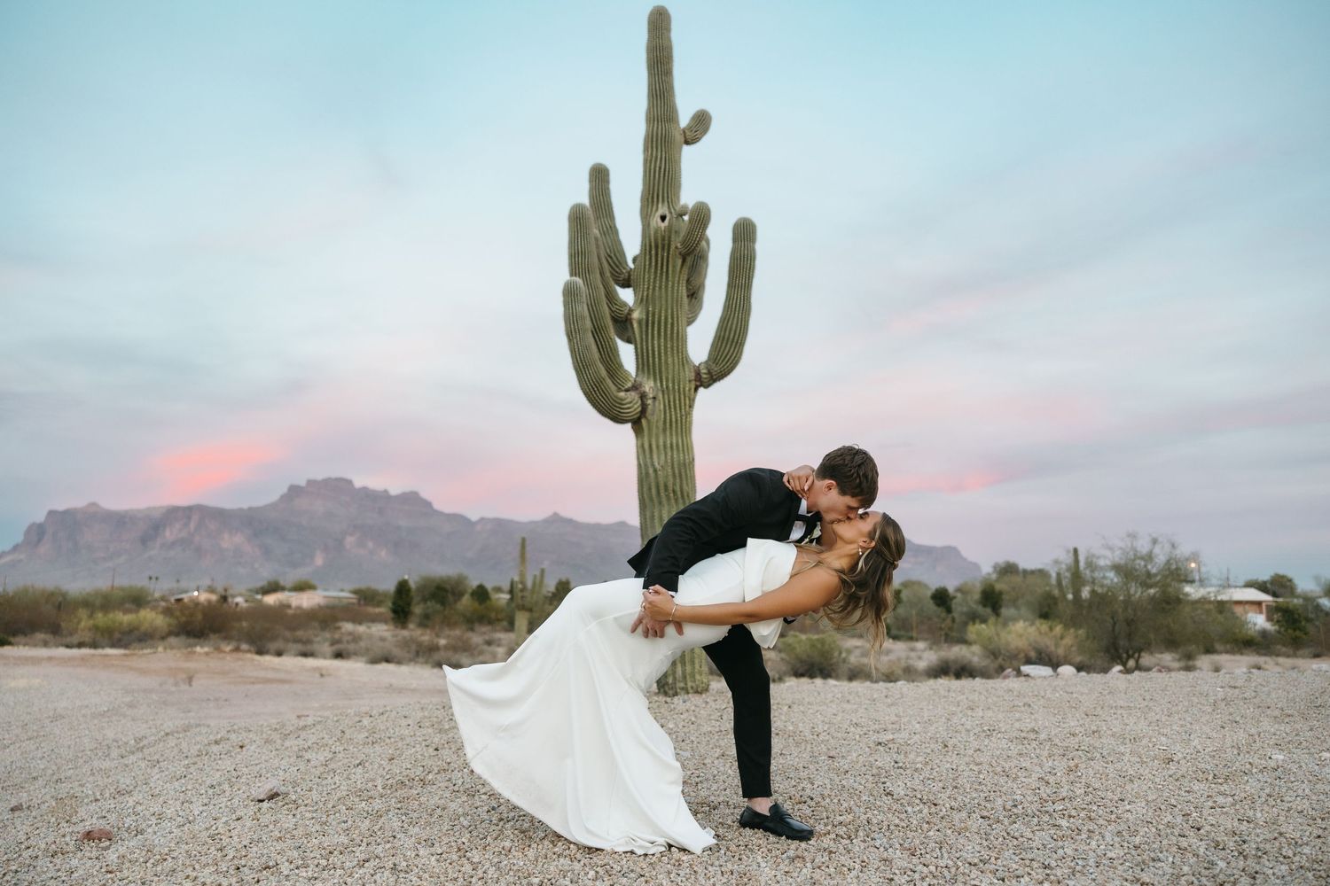 A romantic wedding photo in front of a large saguaro cactus in the desert at sunset.