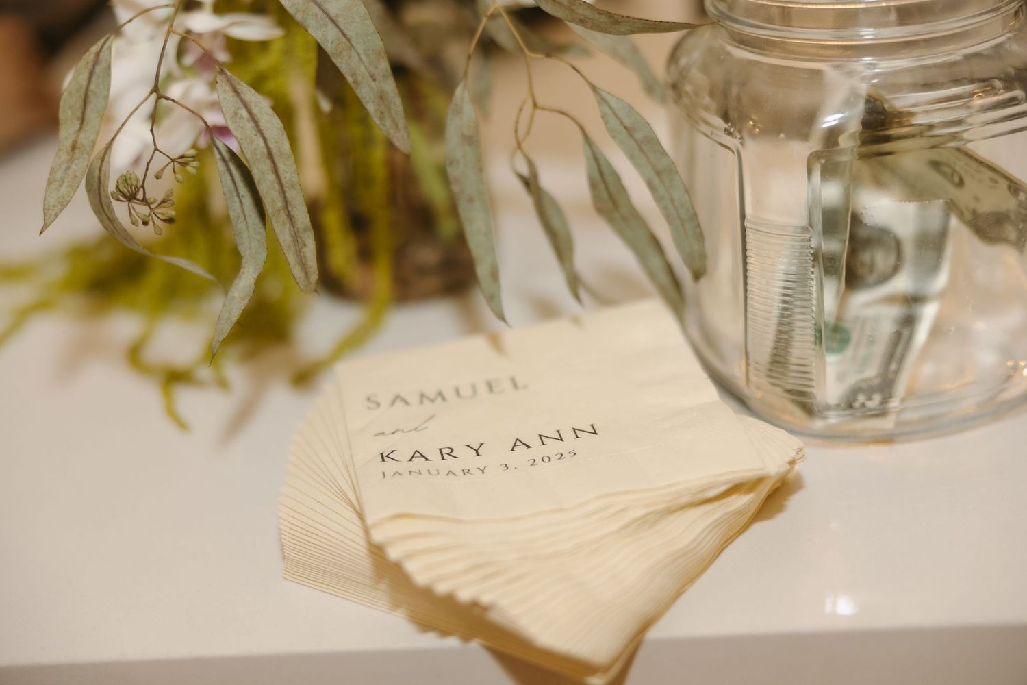 A wedding place card and mason jar sit on a table with greenery decorations.