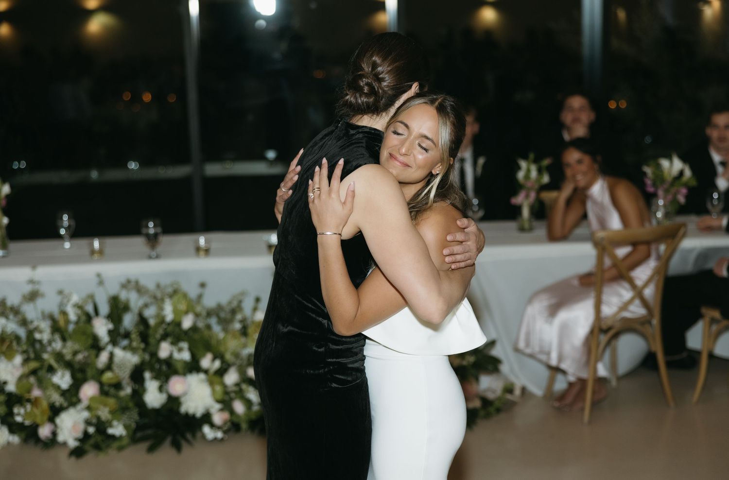 A couple embraces during their wedding dance with floral decorations in the background.