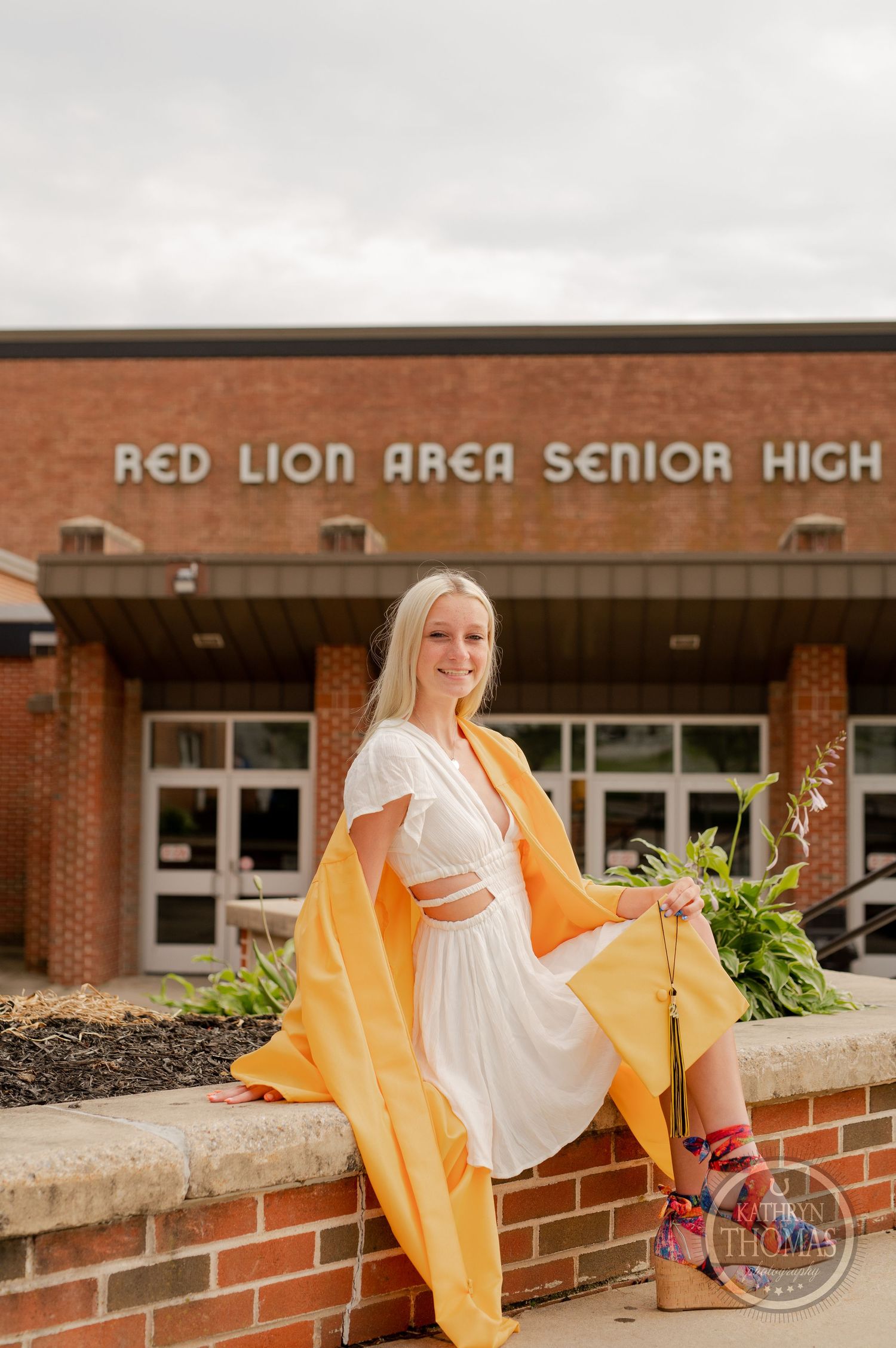 Graduate in yellow gown and white dress poses on brick wall with decorative plants.