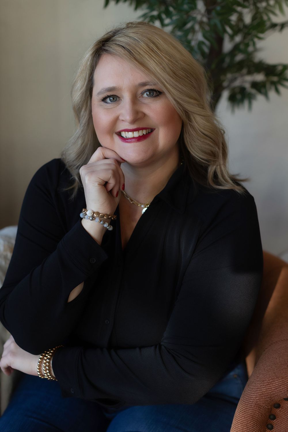 Professional portrait showing a blonde woman in black top wearing gold jewelry seated on a brown chair.