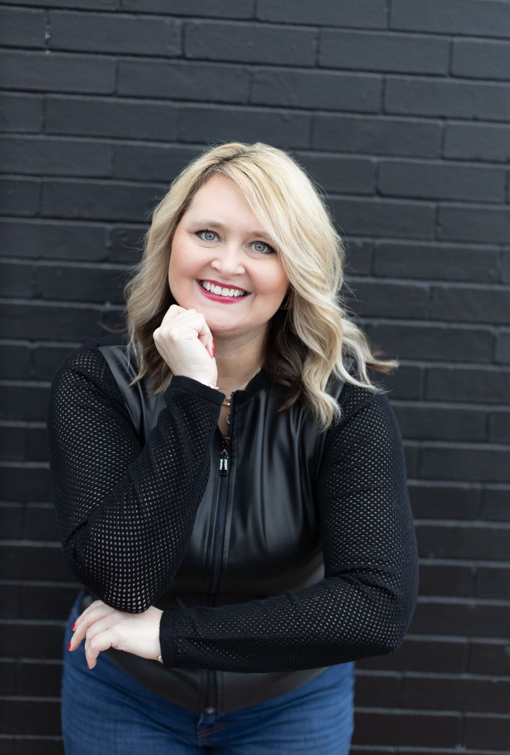 Professional portrait against dark brick wall showing warm smile and stylish black sweater with wavy blonde hair.