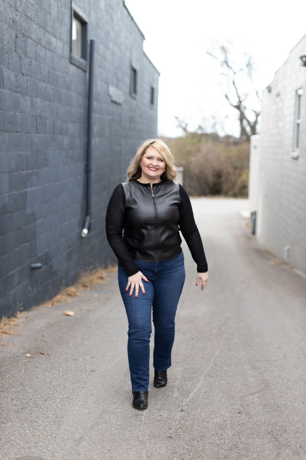 Woman in dark jacket and jeans walking down an alley between gray concrete buildings.