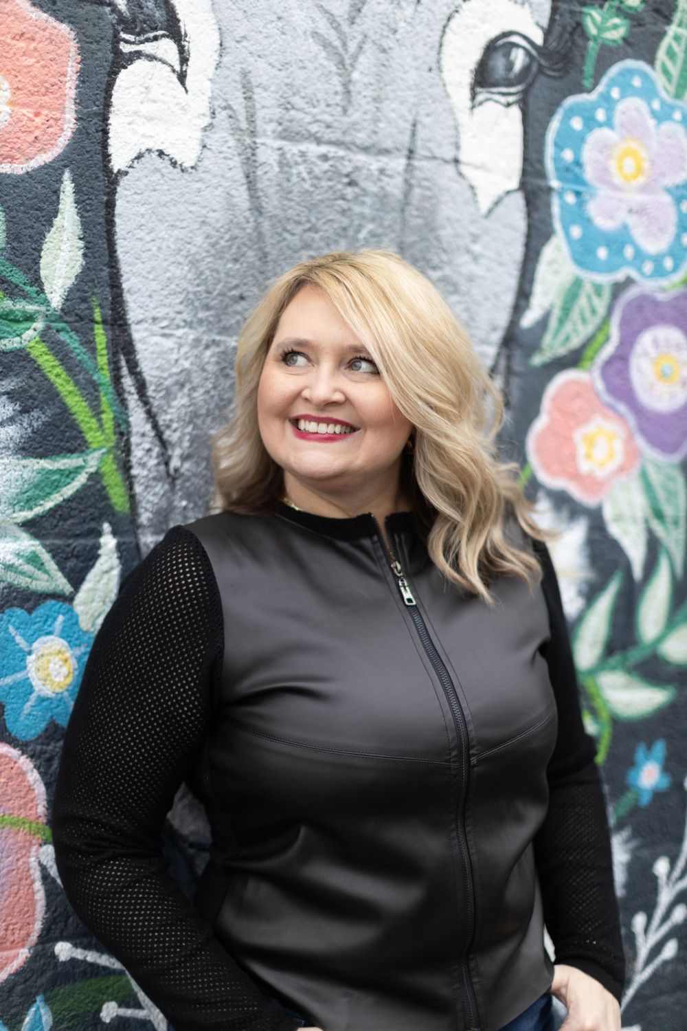 A woman in a black sweater smiles warmly in front of a colorful floral mural on a gray wall.