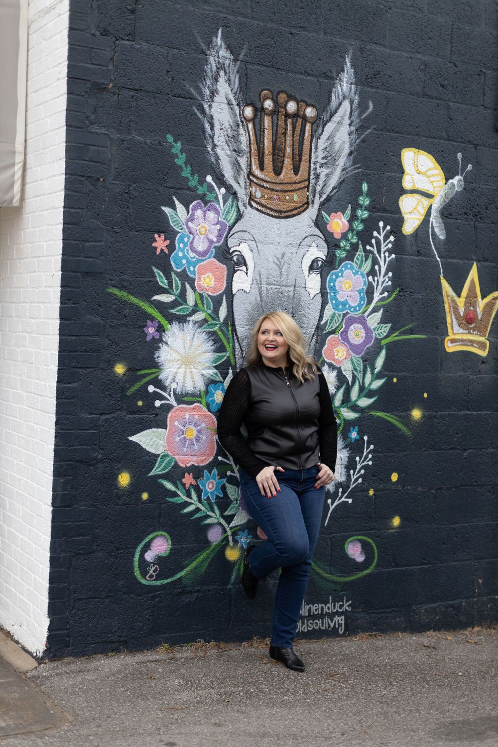 Colorful street art mural featuring floral designs, crowns and wings painted on dark wall with someone posing in front.