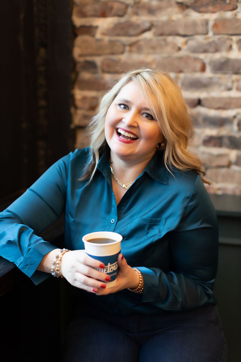 A person in a teal blue blouse enjoys coffee while sitting against a rustic brick wall background.
