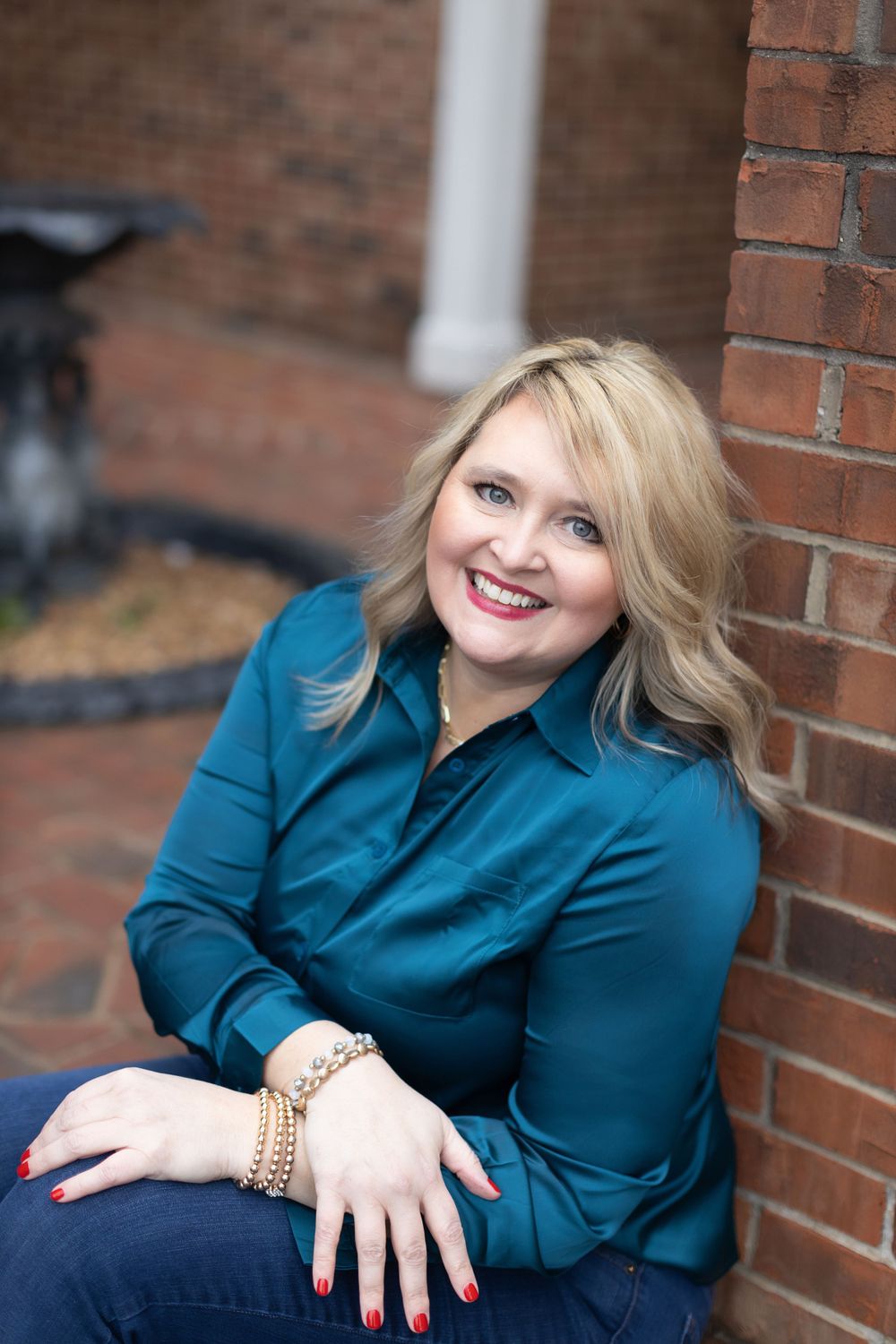 Portrait of a person in teal blouse seated by brick wall wearing gold bracelets and casual attire.