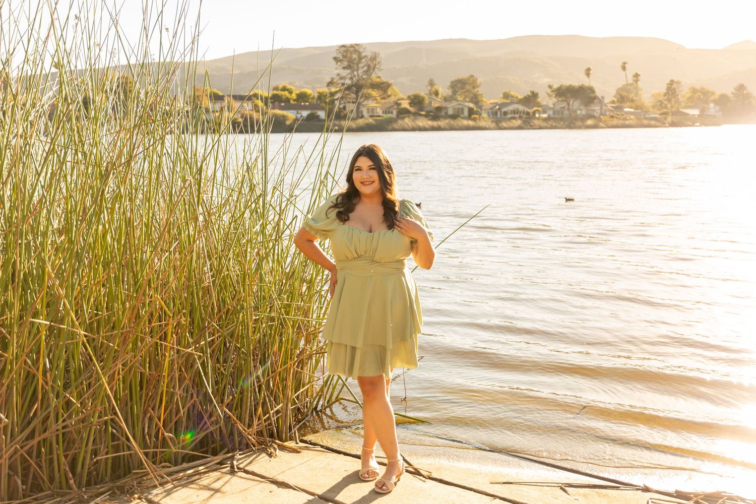 Person in green dress stands among tall reeds at sunset by peaceful lake.
