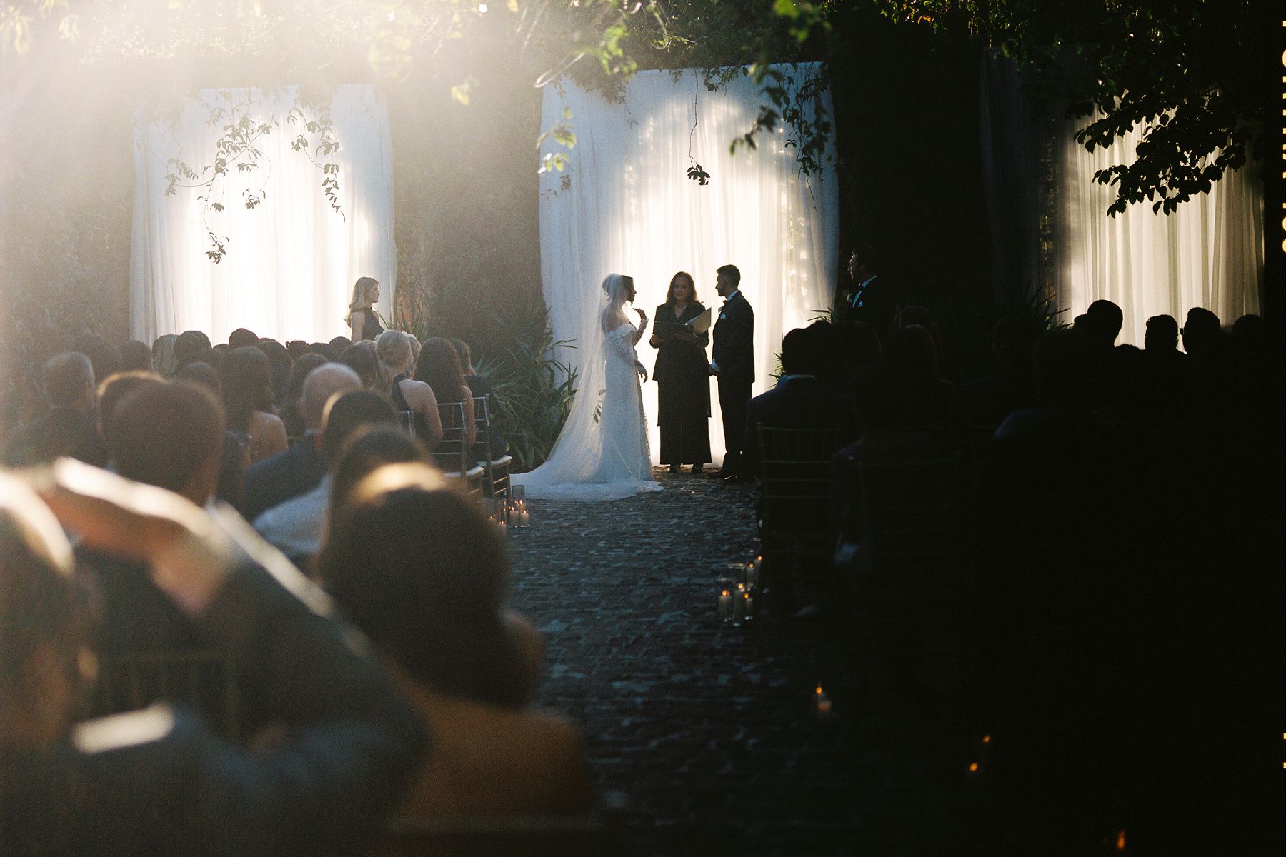 Wedding guests gather in a dimly lit outdoor venue with string lights creating a romantic atmosphere.