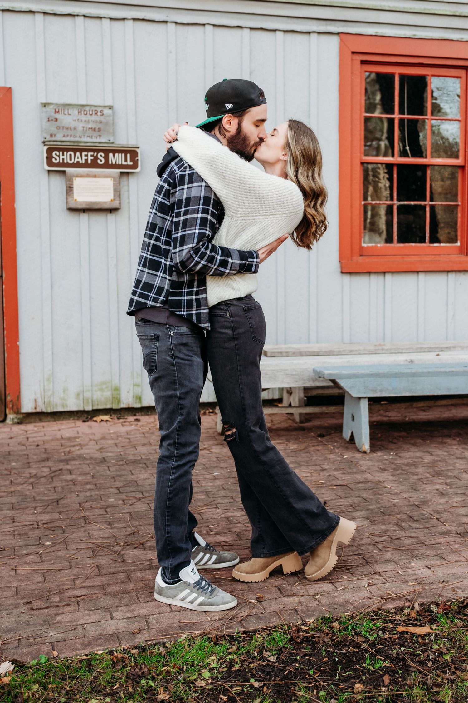 A romantic winter engagement photo outside a rustic Graff's Mill building with red trim and brick path.